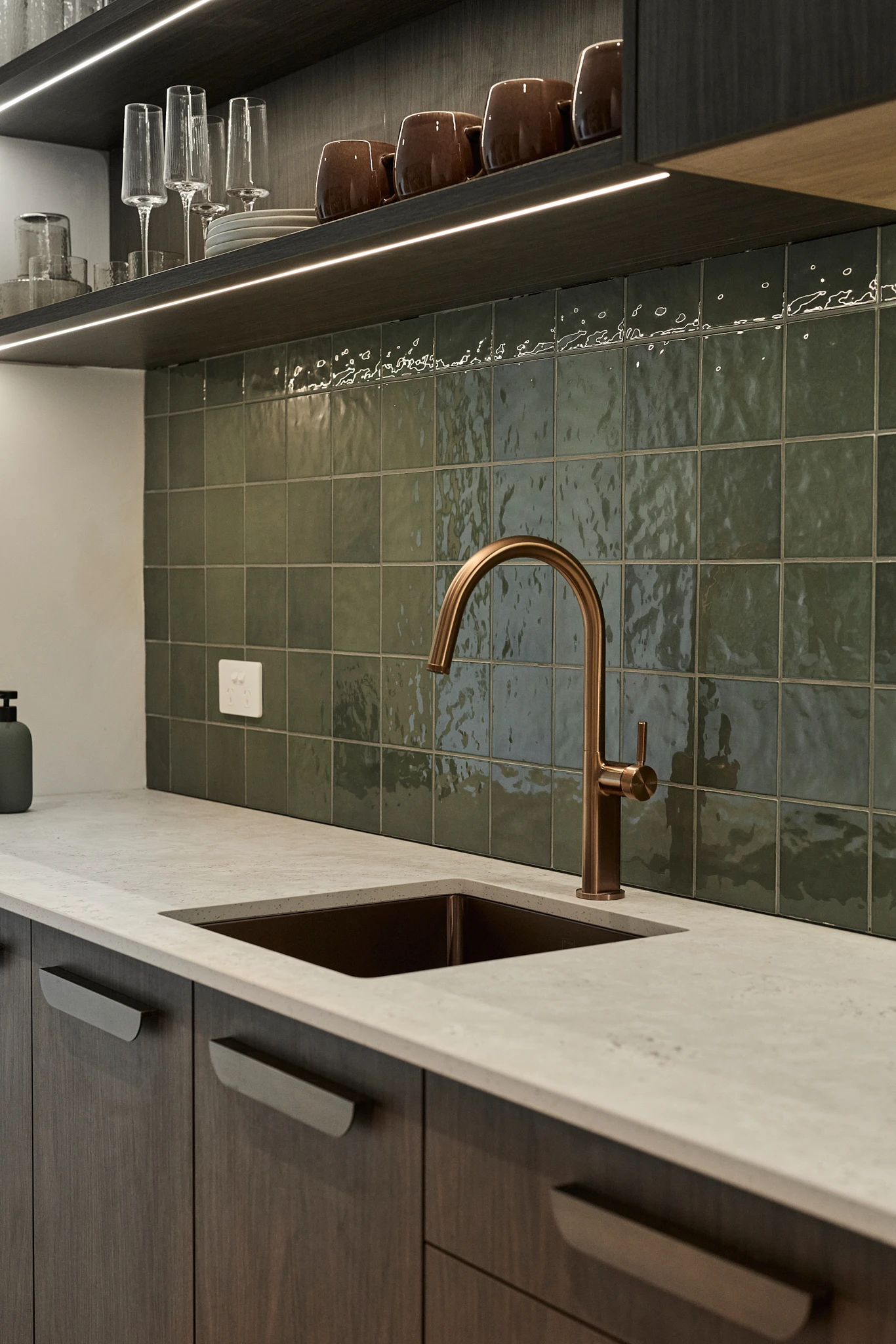 Modern kitchen with green tile backsplash, bronze faucet, white countertop, and open shelving displaying glassware.