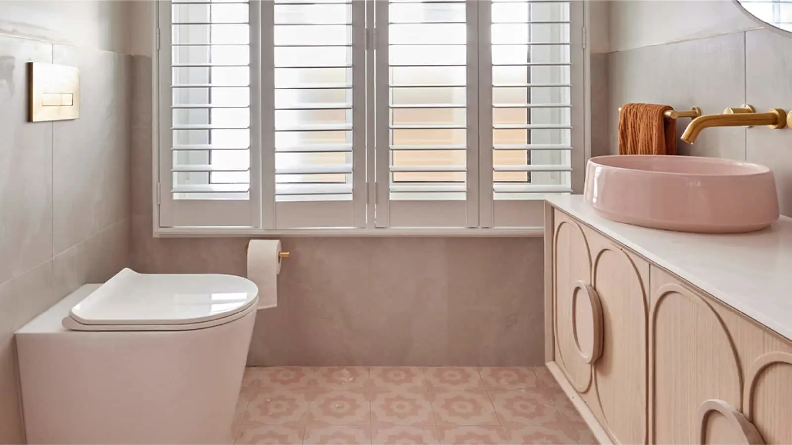 Modern bathroom with white shutters, pink basin, wooden vanity, white toilet, and patterned pink floor tiles.