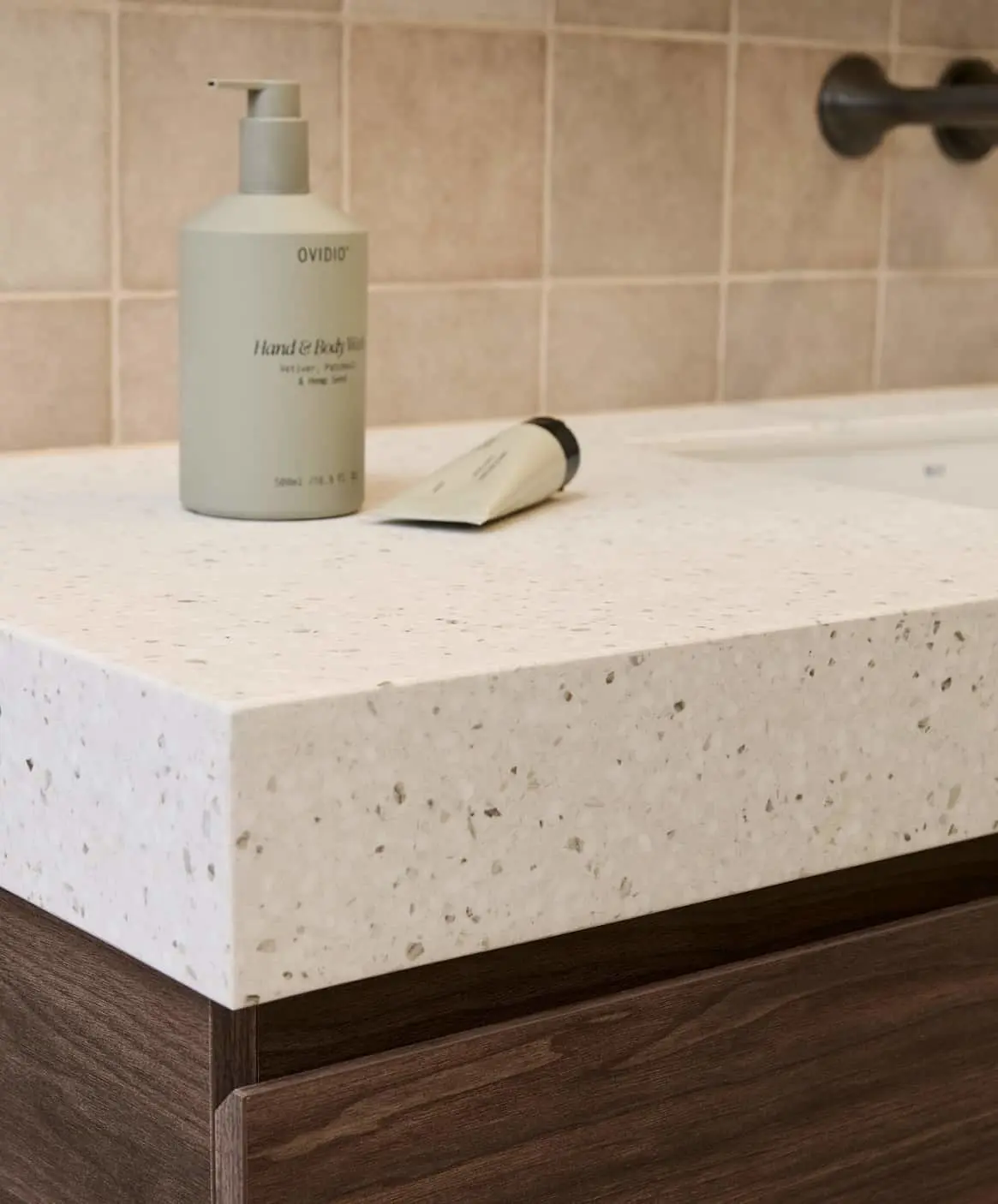 Modern bathroom with terrazzo countertop holding skincare products against beige tiles.