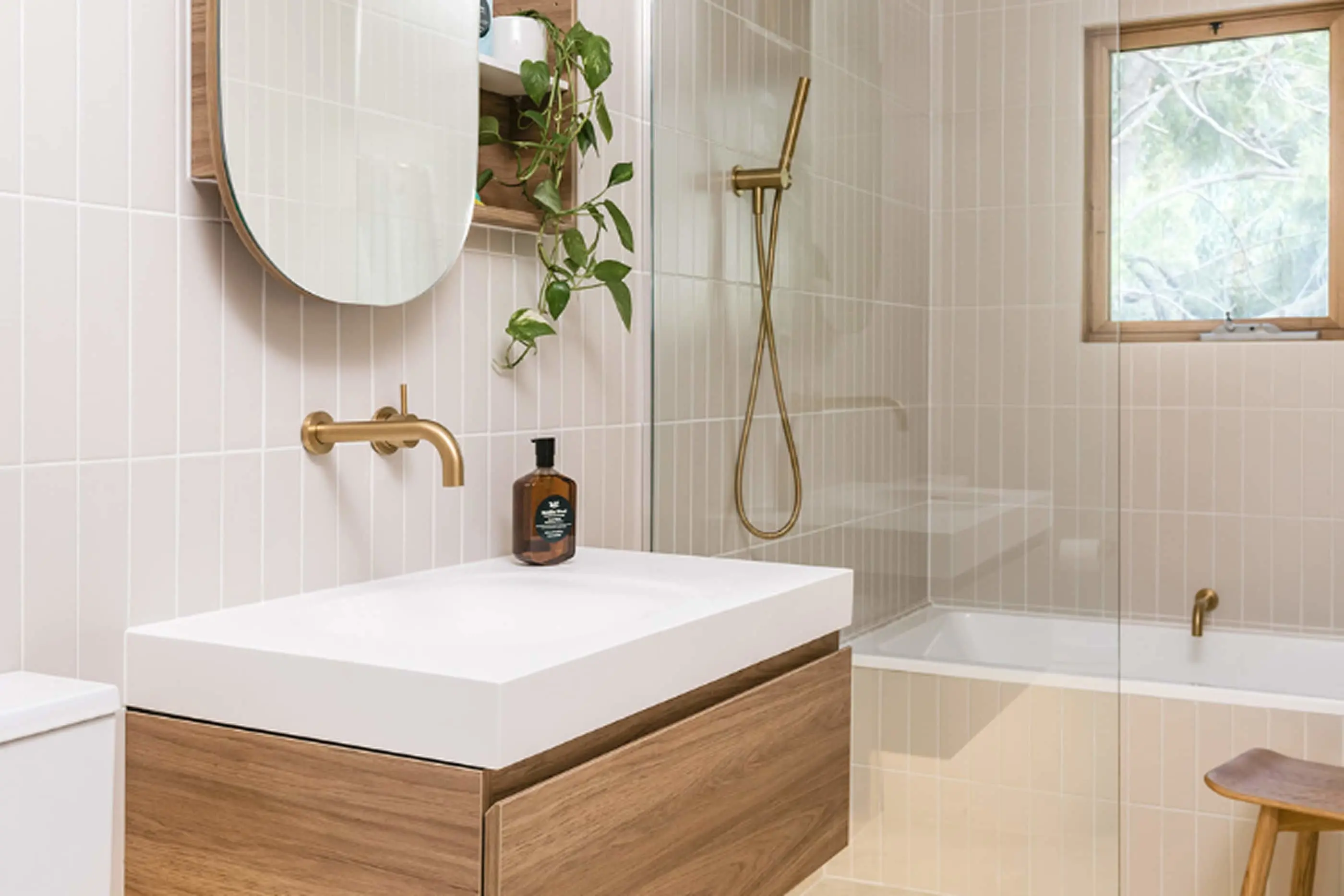 Modern bathroom with white tiles, wooden vanity, gold fixtures, glass shower, and potted plant near circular mirror.