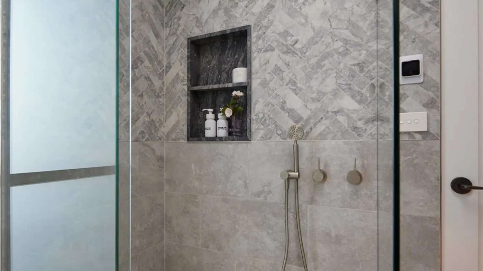 Modern shower with marble herringbone tile, built-in niche holding toiletries and flowers, and handheld showerhead.