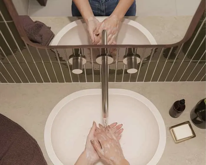 Person washing hands with soap in a bathroom sink with modern faucet, viewed from above through a mirror.