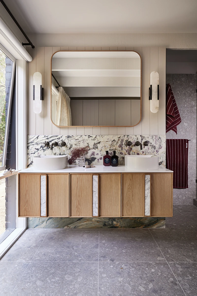 Modern bathroom with wooden vanity, marble backsplash, round vessel sinks, and gold-framed mirror flanked by wall sconces.