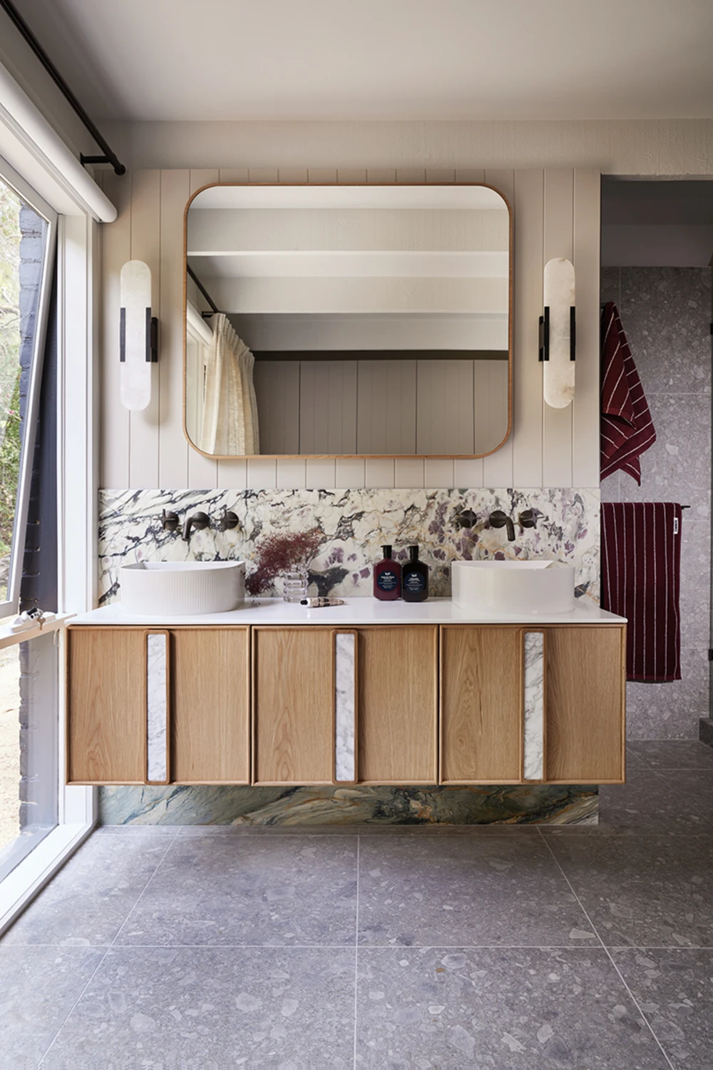 Modern bathroom with wooden vanity, marble backsplash, round vessel sinks, and gold-framed mirror flanked by wall sconces.