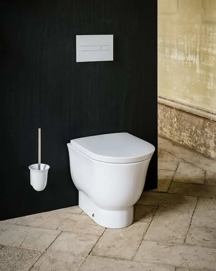 Modern white toilet against black wall with flush panel and toilet brush on stone tile floor.