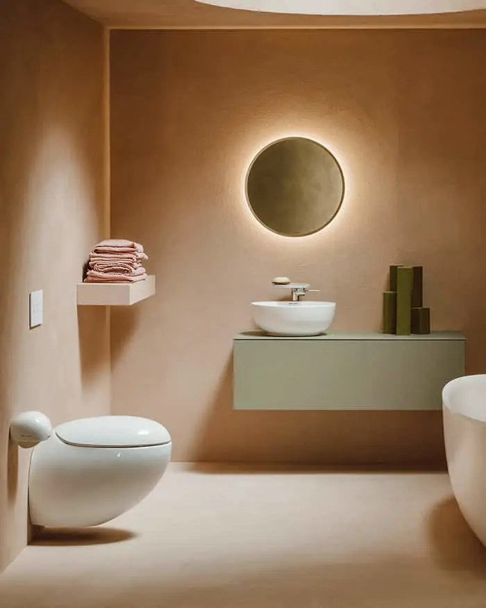 Bathroom with terracotta walls, white wall-hung toilet, green floating vanity with vessel sink, round backlit mirror, and pink towels on a shelf.