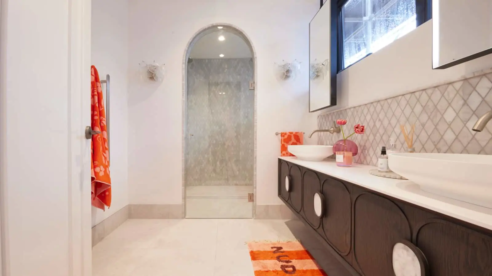 Modern bathroom with arched glass shower door, dark vanity, white sink, orange towels and rug, and pink floral accents.