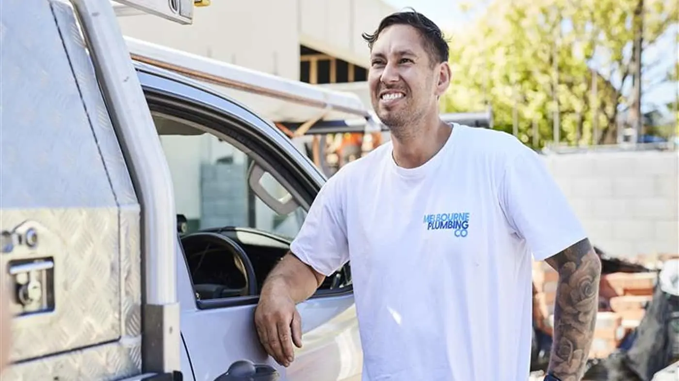 Plumber in white Melbourne Plumbing Co shirt smiling next to work van on sunny day, with tattooed arm visible.