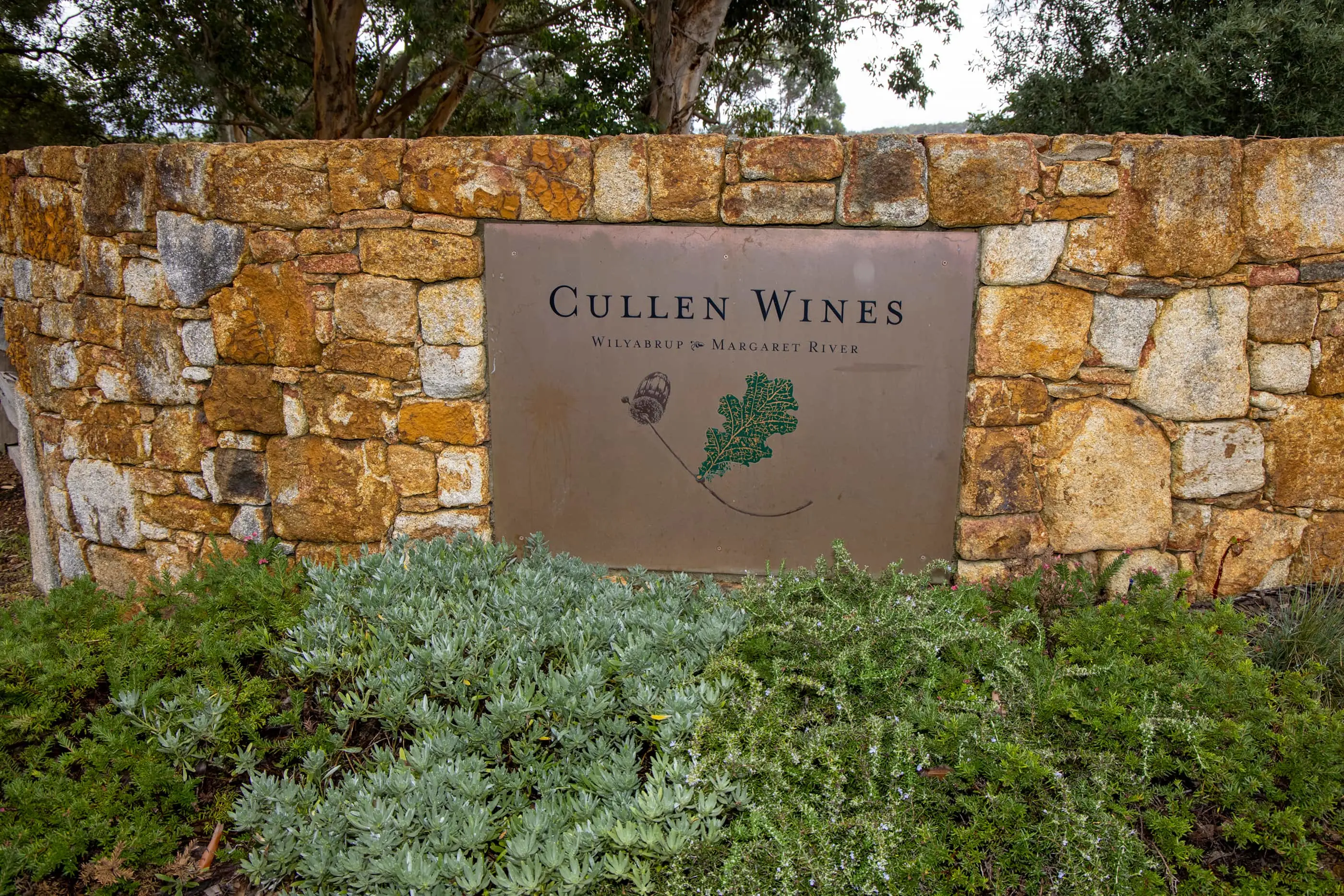 Stone wall entrance sign for Cullen Wines in Wilyabrup, Margaret River, with green shrubs at the base and eucalyptus trees behind.