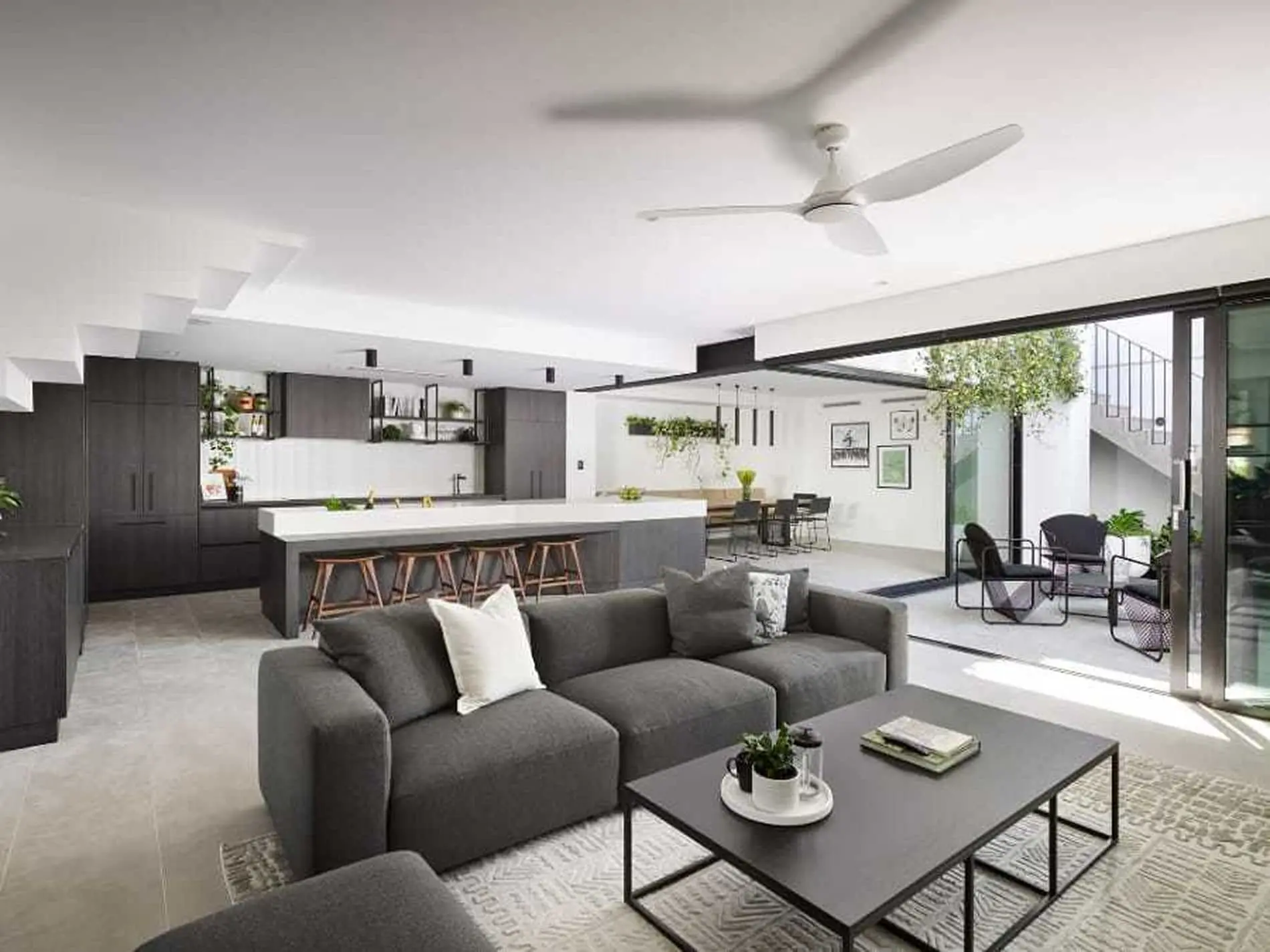 Modern open-concept living space with gray sofa, kitchen island with bar stools, and sliding doors to outdoor area with plants.