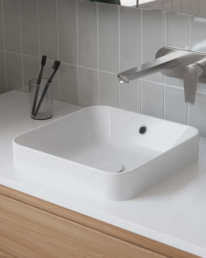 White square vessel sink on a white countertop with a wall-mounted silver faucet, toothbrushes in a cup, and grey tile wall.