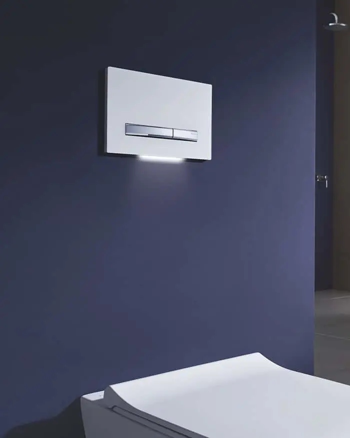 Modern white toilet flush panel with illumination mounted on dark blue bathroom wall above white toilet fixture.