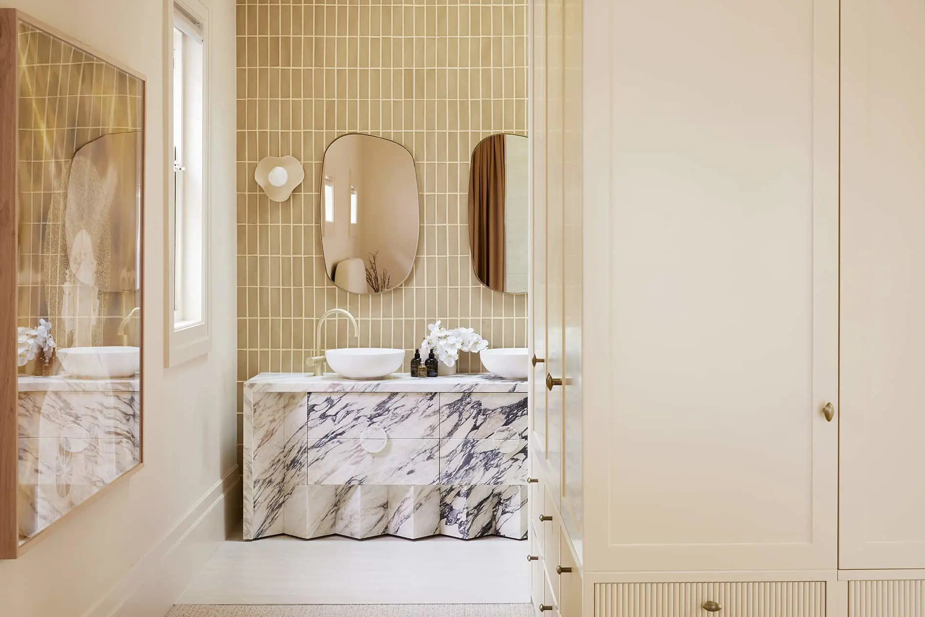 Modern bathroom with marble vanity, two white vessel sinks, oval mirrors, and beige tile wall accent.