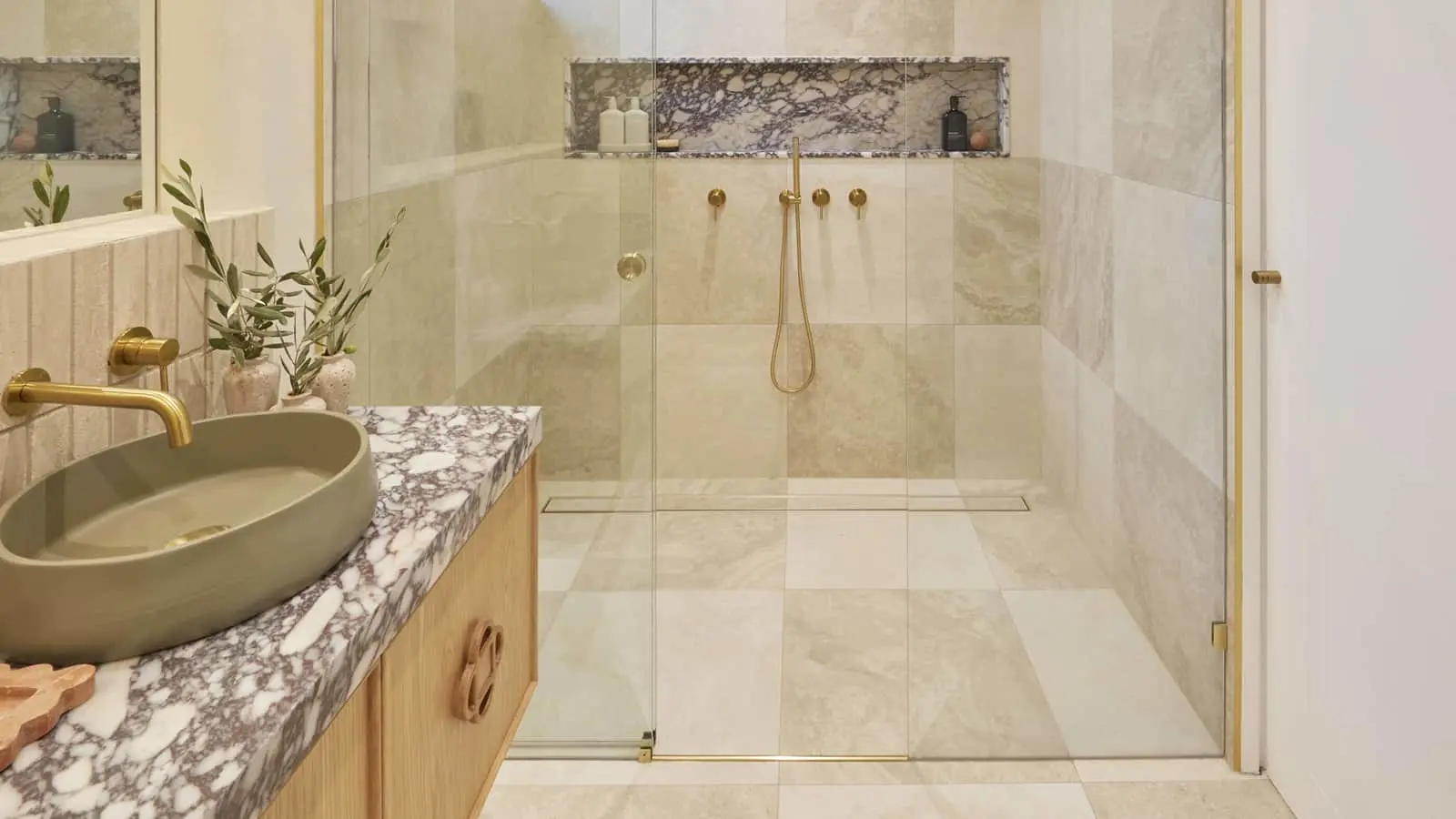 Modern bathroom with glass shower, beige marble tiles, green sink on marble countertop, and gold fixtures.