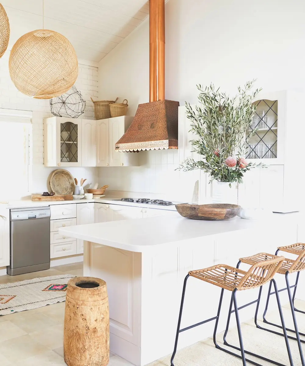 Bright white kitchen with copper range hood, wicker pendant light, rattan bar stools, and olive plant on island counter.