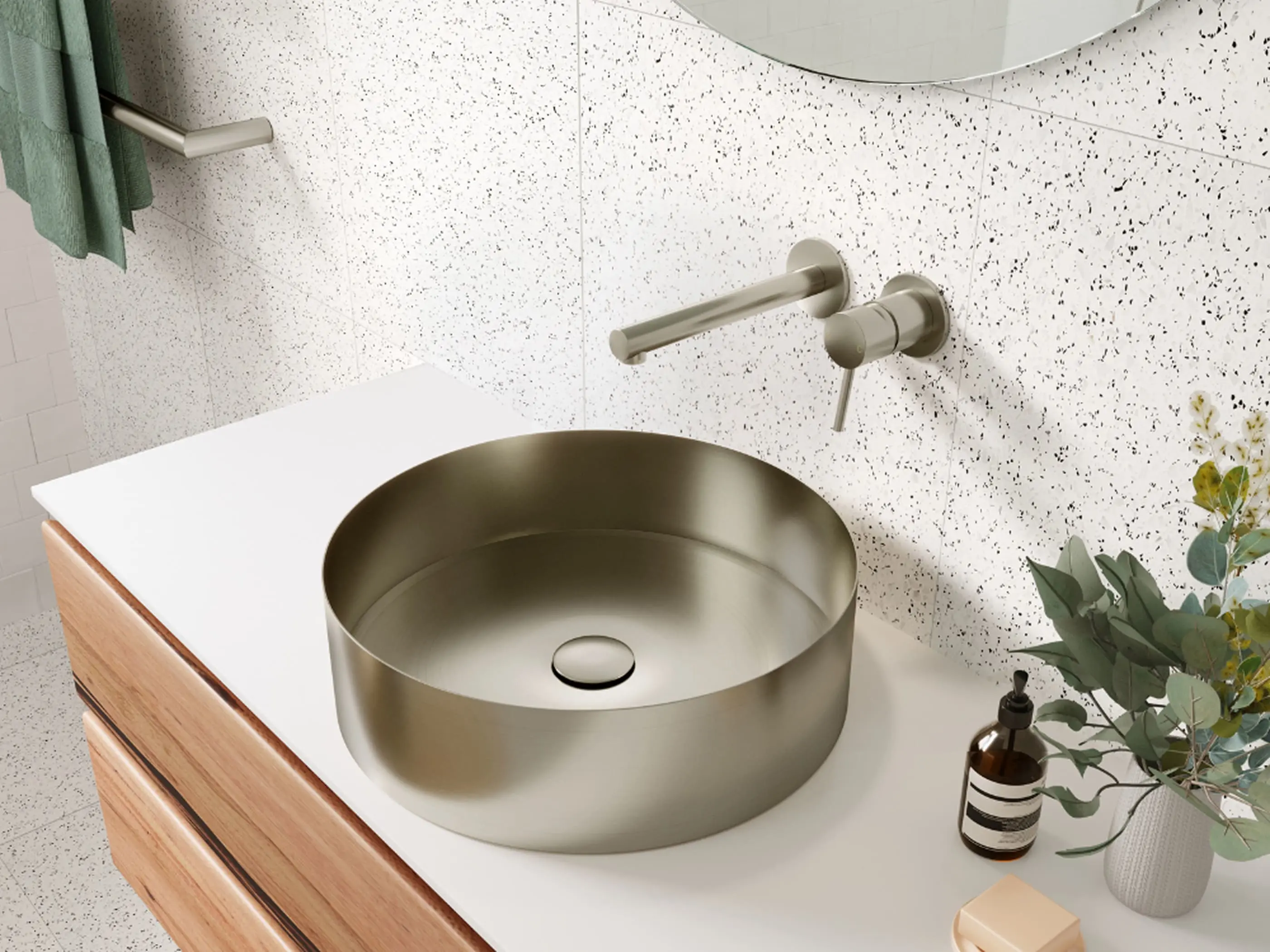 Modern bathroom with round brushed metal sink, wall-mounted faucet, wooden vanity, and decorative plants on white counter.