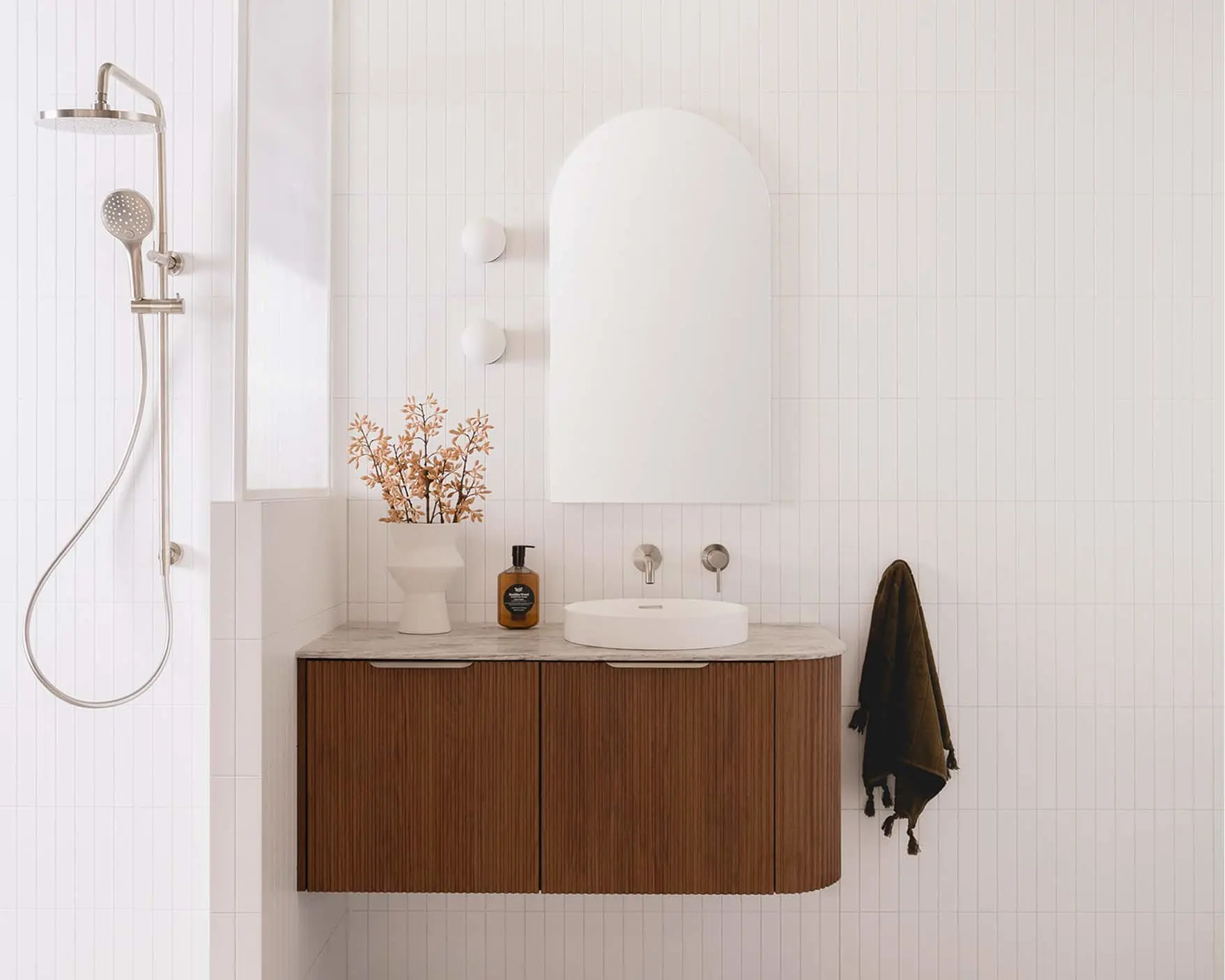 Modern white bathroom with wooden vanity, arched mirror, rainfall shower, and decorative dried plant in a minimalist design.