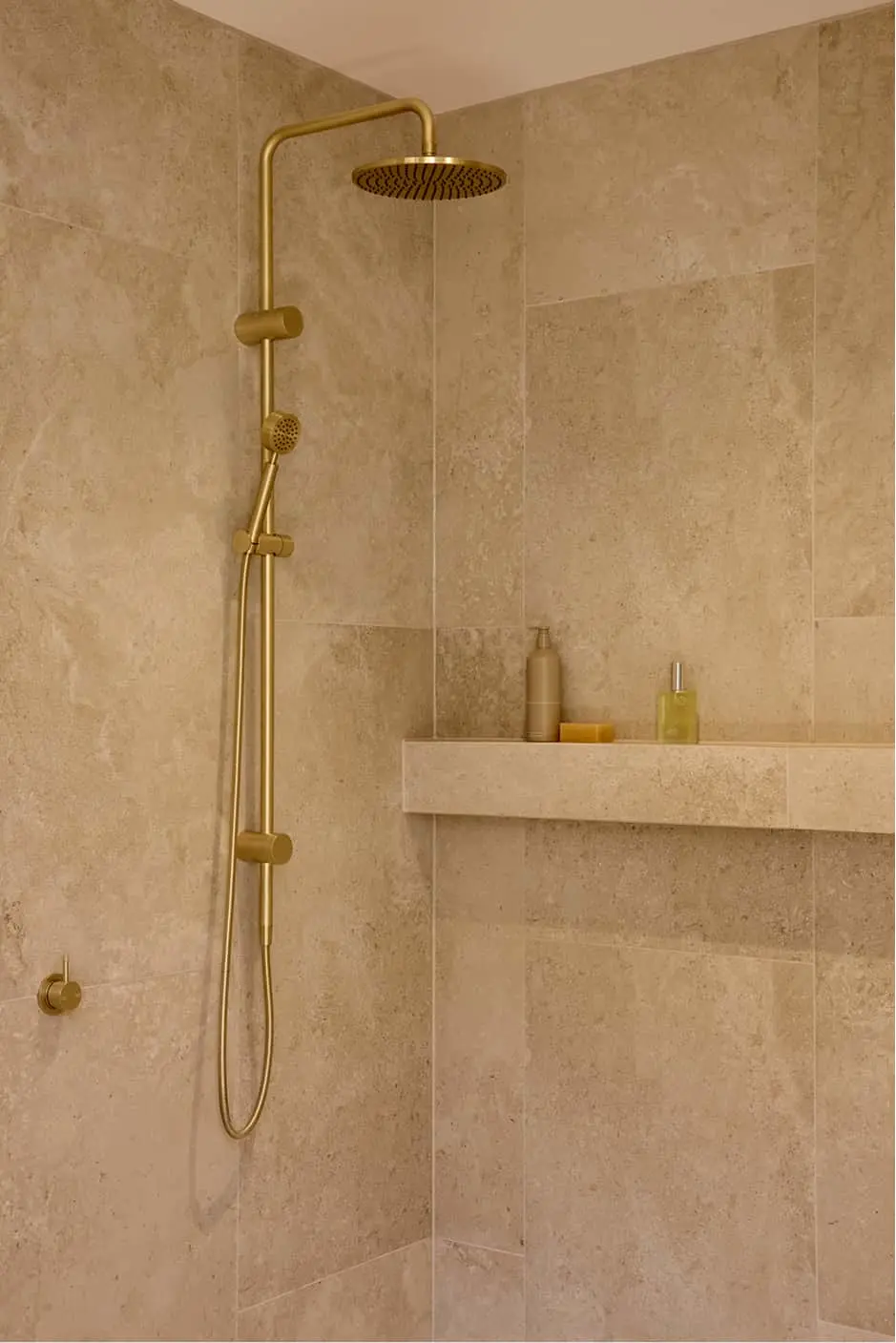 Walk-in shower with beige stone tiles, gold rainfall showerhead, handheld attachment, and a built-in shelf with toiletries.