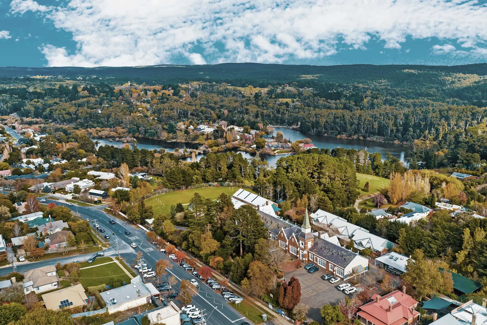 Aerial view of a small town with winding river, autumn trees, church spire, and mountains in the background under blue sky.