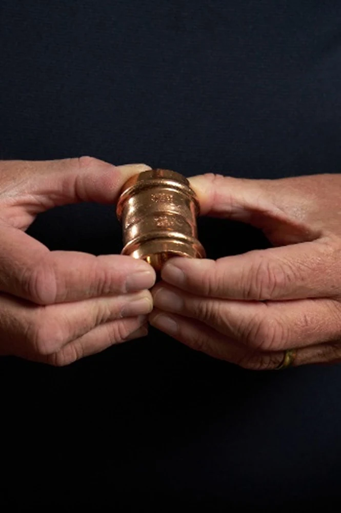 Hands holding a brass or copper plumbing fitting against a dark background.