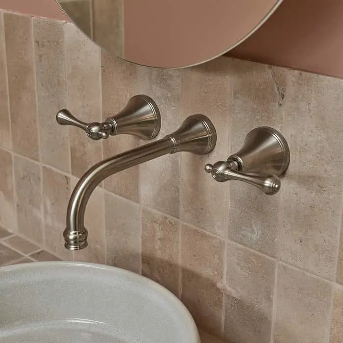 Wall-mounted brushed nickel bathroom faucet with two handles above a white sink, against beige tiled wall.
