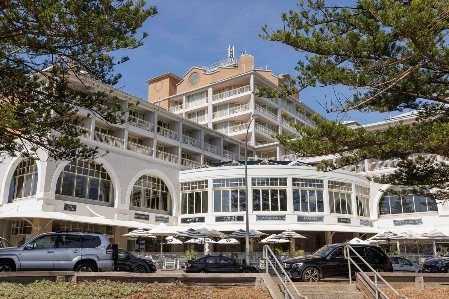 Beachfront hotel with white arched storefronts, outdoor dining umbrellas, and parked cars in front under pine trees.
