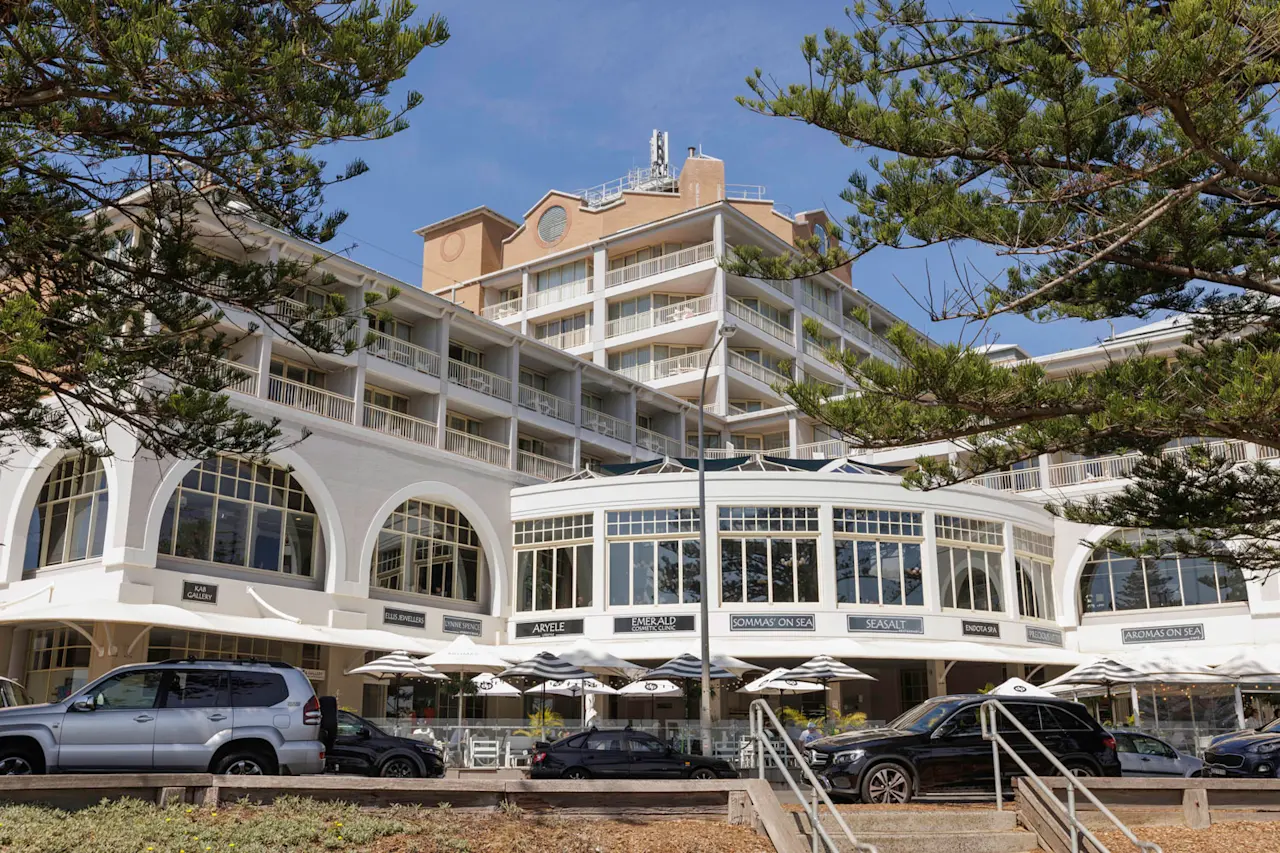 Beachfront hotel with white arched storefronts, outdoor dining umbrellas, and parked cars in front under pine trees.