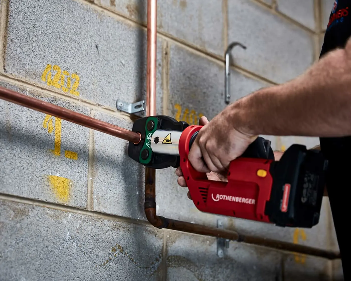 Hand using red Rothenberger power tool to connect copper pipes on concrete block wall with yellow markings.