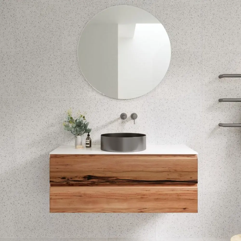 Modern bathroom vanity with wooden cabinet, white countertop, black vessel sink, round mirror, and small plant.
