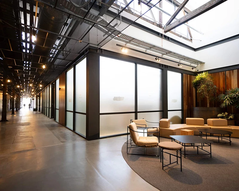 Modern interior with a long corridor, glass partition walls, and a seating area with tan chairs and low tables.