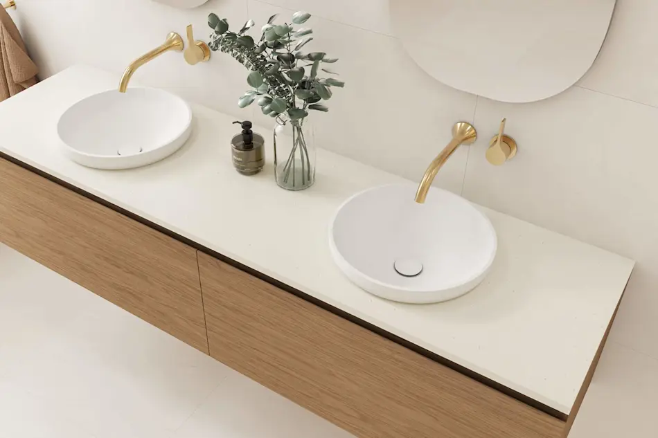 Modern bathroom vanity with dual white vessel sinks, brass faucets, wooden cabinet, and eucalyptus in glass vase.