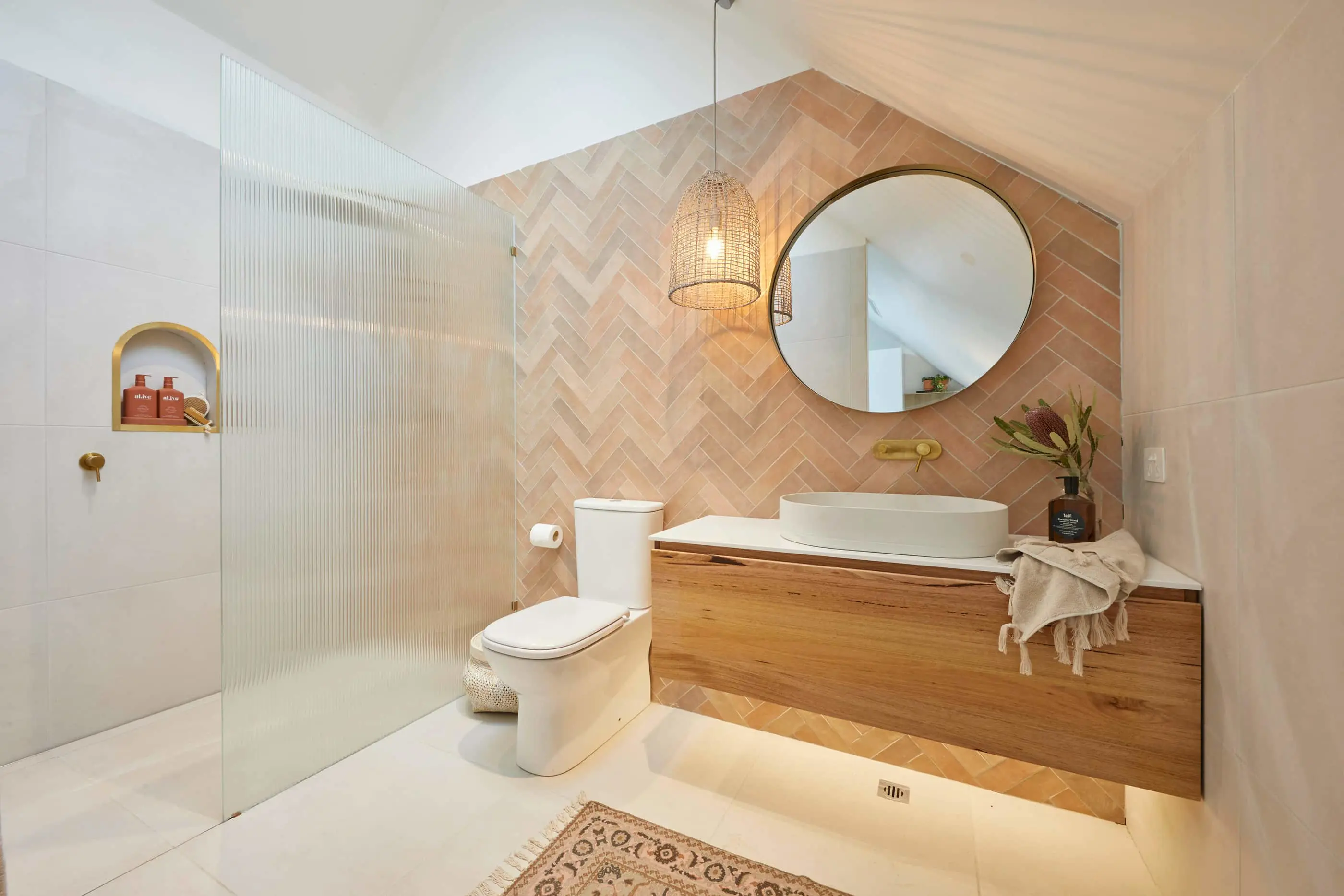 Modern bathroom with peach herringbone tile, wooden vanity, round mirror, wicker pendant light, and glass shower partition.