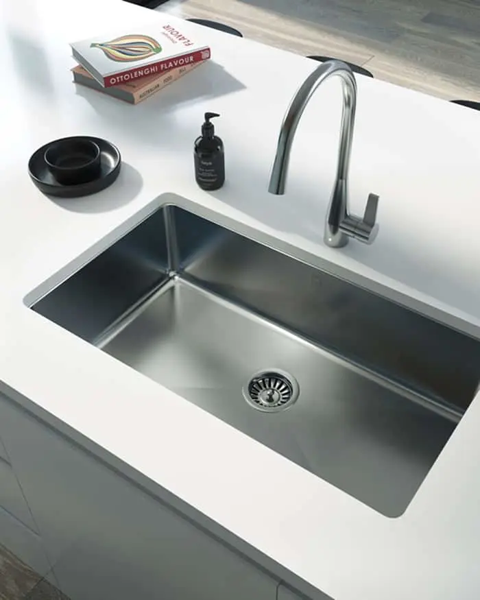 Modern stainless steel kitchen sink with curved faucet, soap dispenser, black dish, and cookbook on white countertop.