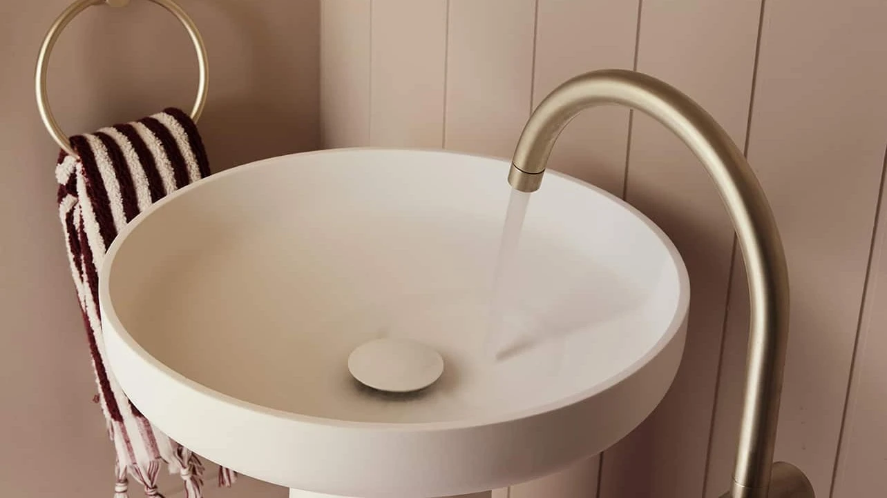 Modern bathroom with white pedestal sink, brass faucet, striped hand towel, and partial round mirror on beige walls.