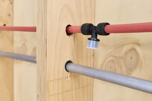 Red and gray PEX plumbing pipes running through wooden wall studs with a black elbow fitting connector.