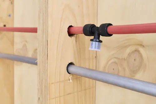 Red and gray PEX plumbing pipes running through wooden wall studs with a black elbow fitting connector.