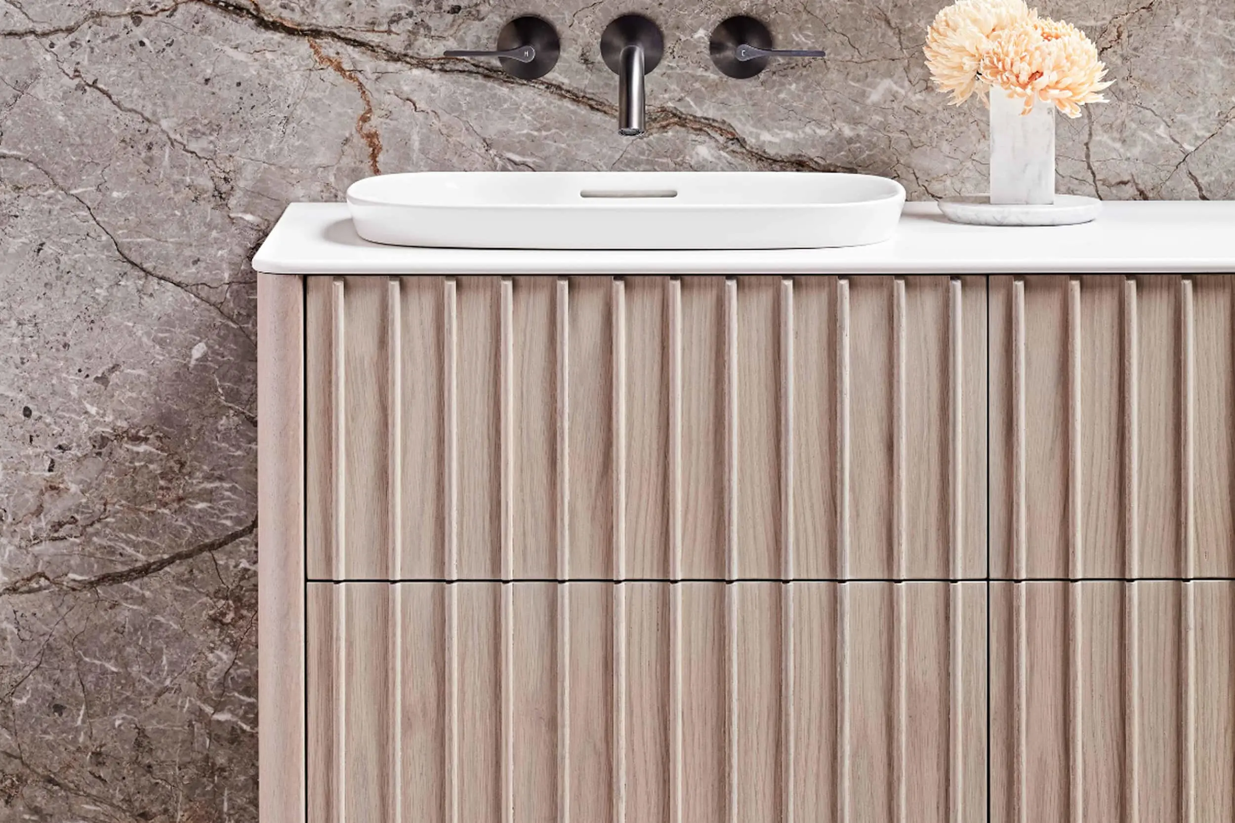 Modern bathroom vanity with white oval sink, wooden ribbed cabinet, marble wall, and peach flower in small vase.