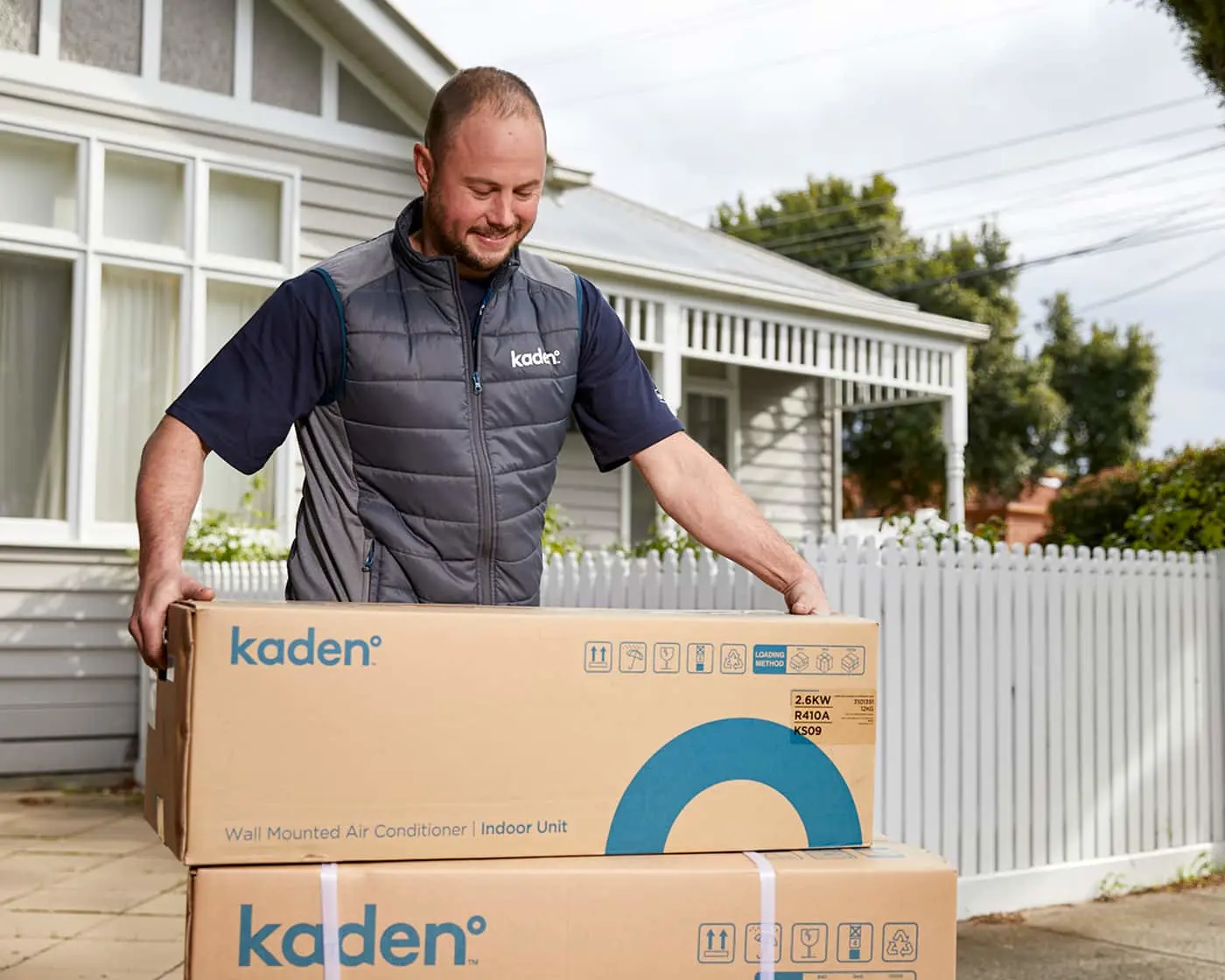 Person in gray Kaden vest delivering air conditioner units outside a white house with picket fence.