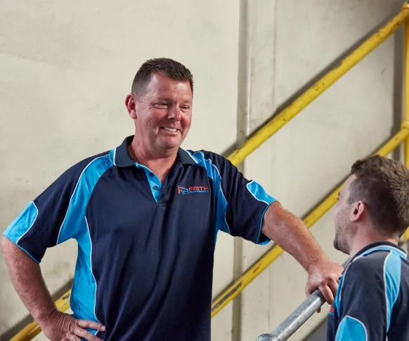 Two workers in blue and black uniforms conversing near yellow railings in an industrial setting.