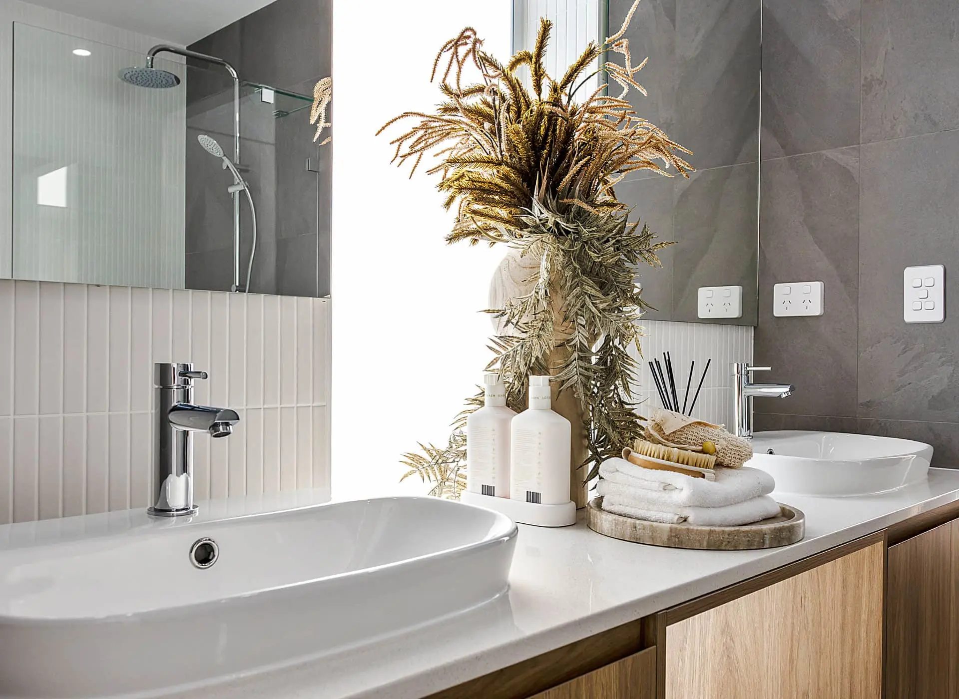Modern bathroom with white sinks, dried floral arrangement, toiletries on wooden tray, and glass shower enclosure.