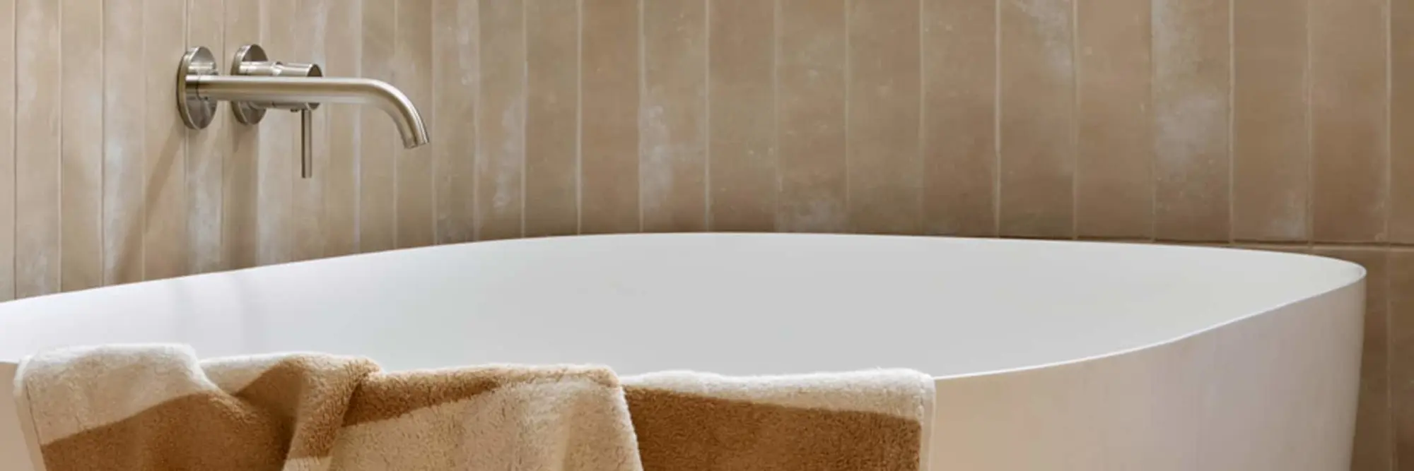 Modern white freestanding bathtub with wall-mounted stainless steel faucet against beige striped tile wall.