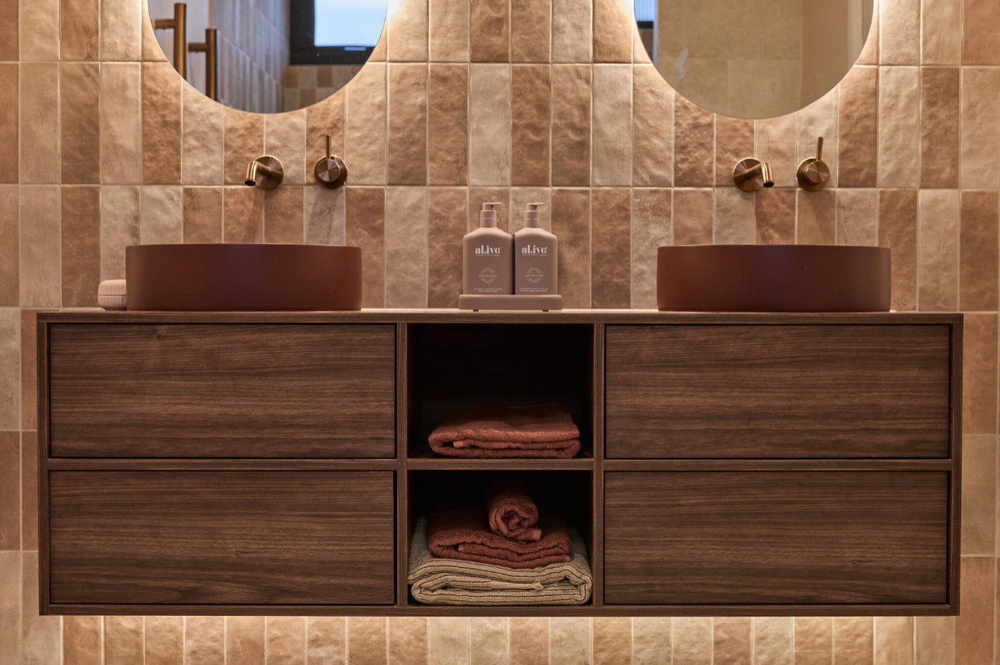 Modern bathroom with dual brown vessel sinks, wooden vanity, beige tiled walls, and neatly stacked towels.