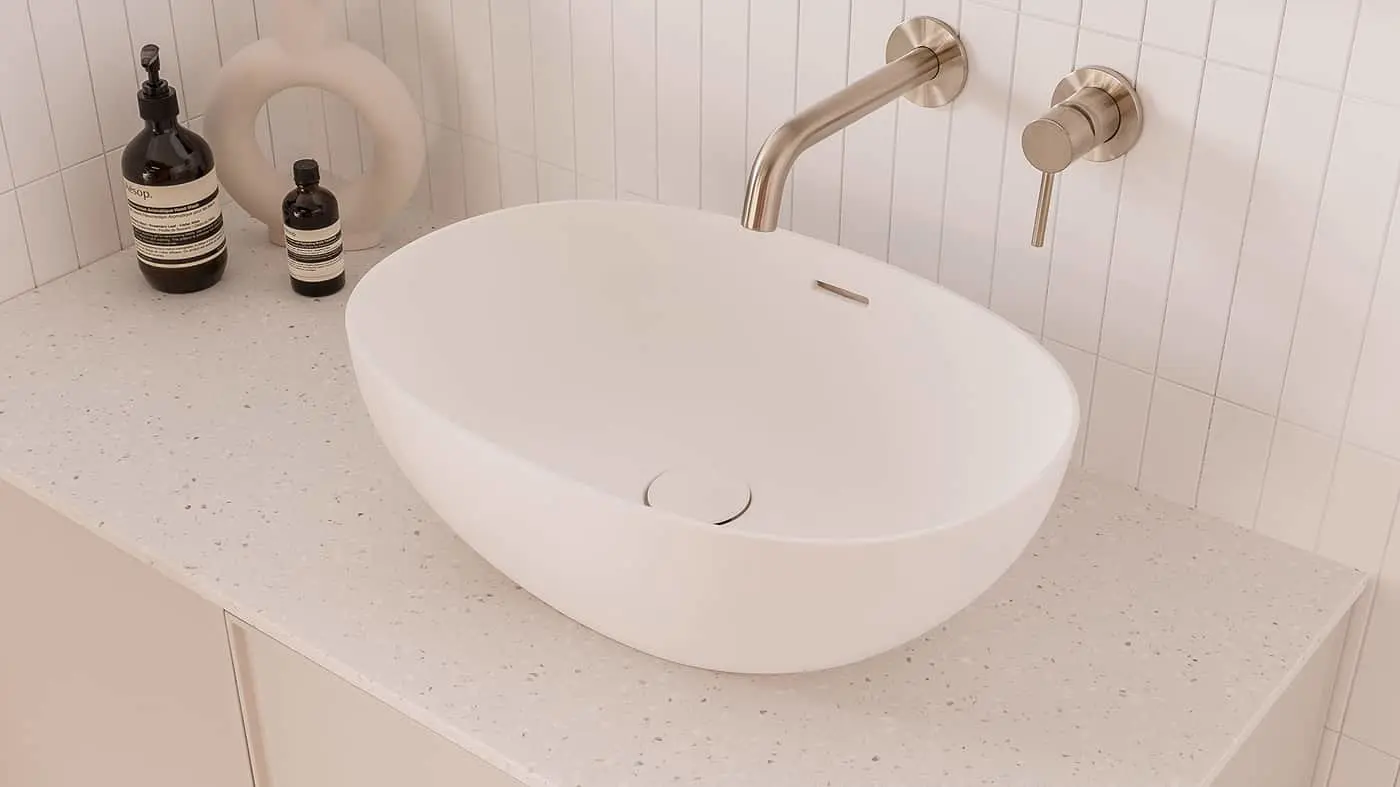 Modern white oval bathroom sink with brass wall-mounted faucet, terrazzo countertop, and dark glass soap bottles.