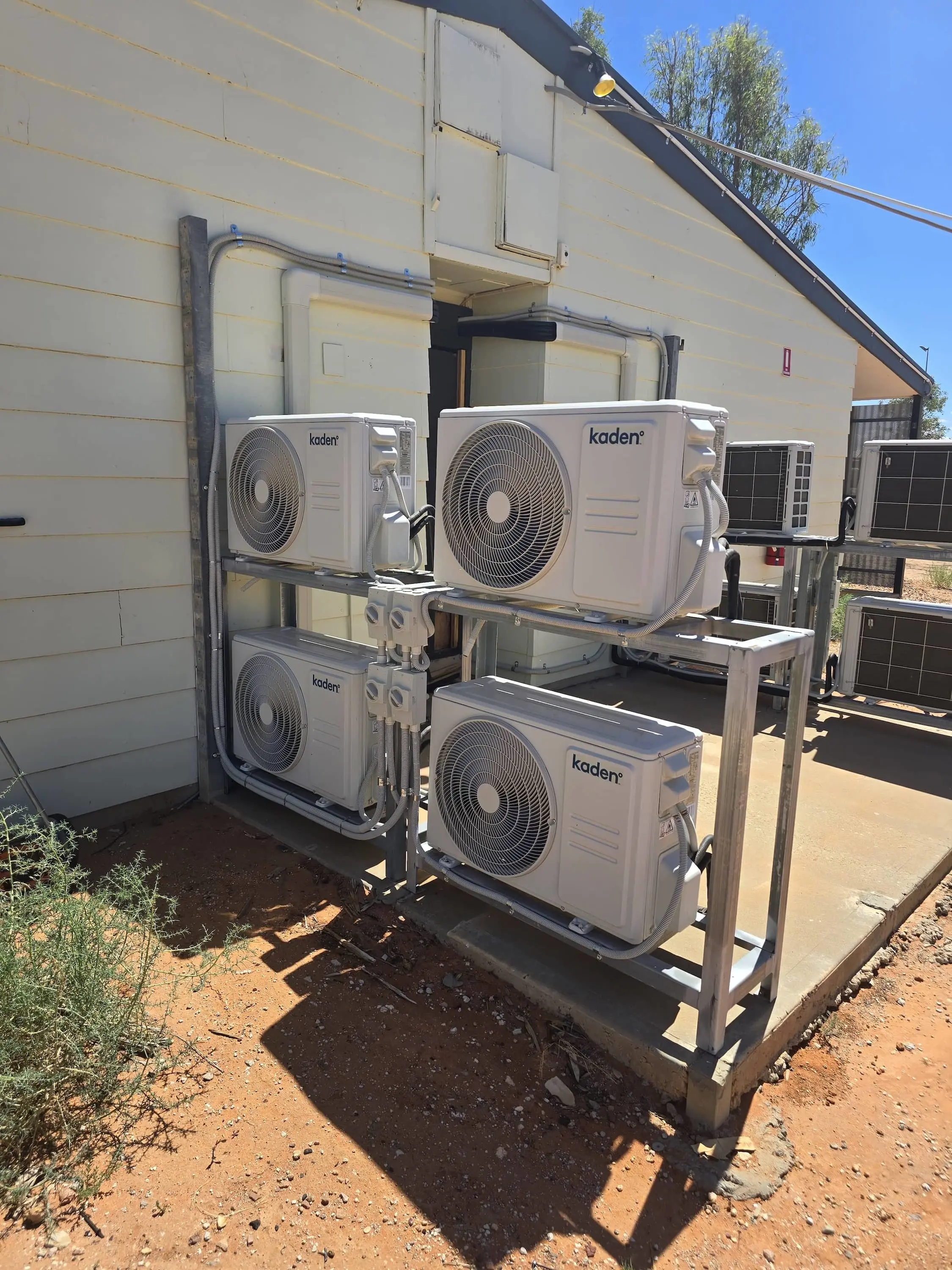 Four Kaden air conditioning outdoor units mounted on metal frame against white building exterior on concrete pad.