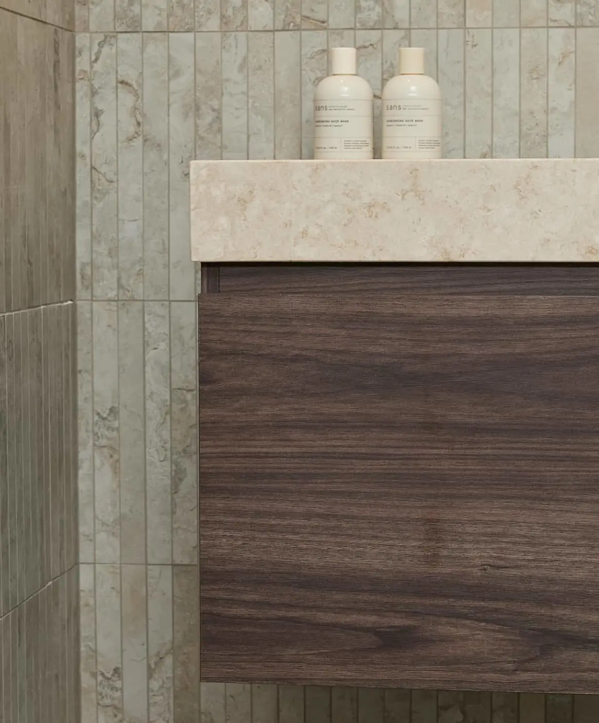 Two white shampoo bottles on a stone countertop with dark wood cabinetry and light marble-patterned wall tiles.
