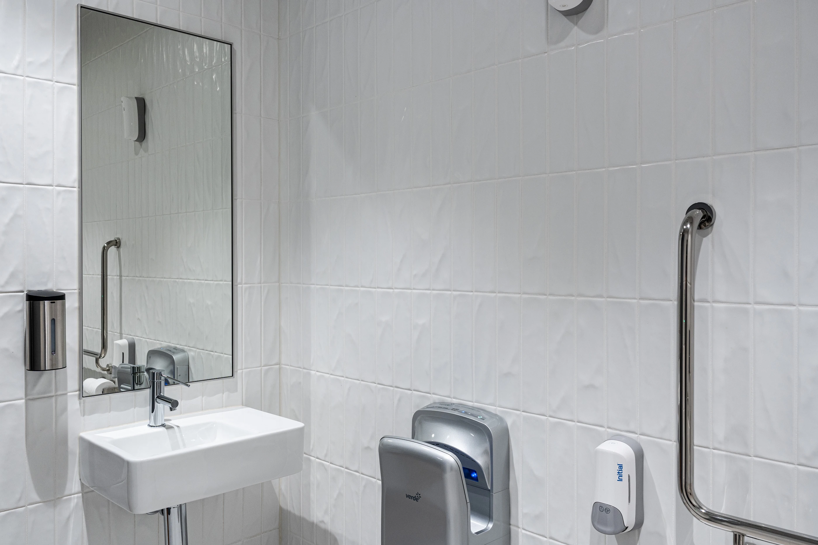 Modern white bathroom with sink, mirror, hand dryer, soap dispenser, and safety grab bar on tiled walls.