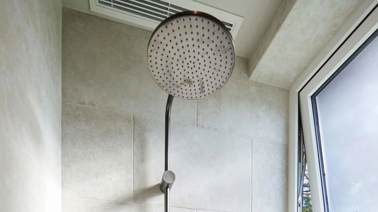 Large circular rainfall showerhead mounted on a metal arm in a modern bathroom with light stone tile walls.