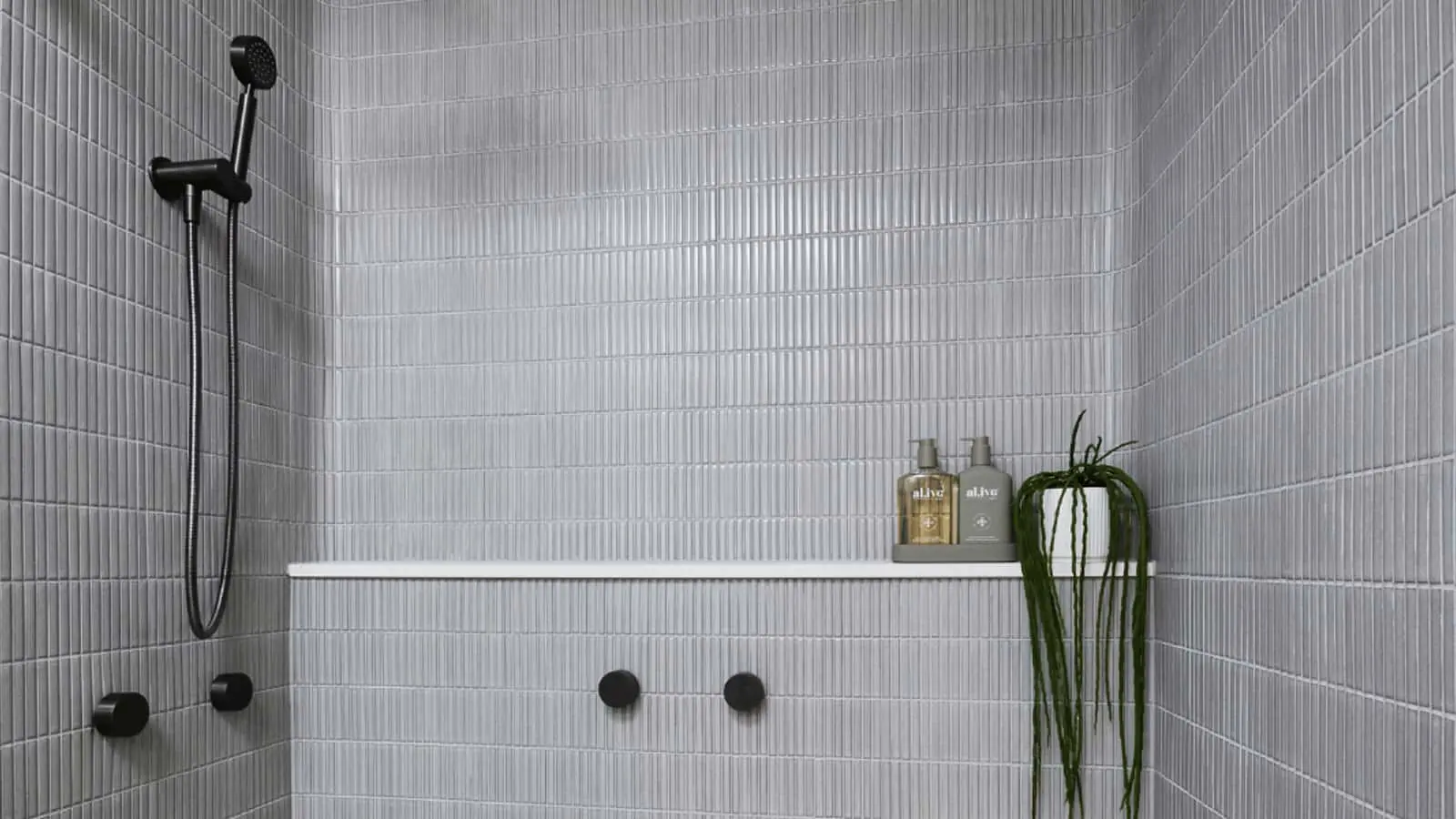 Modern shower with light gray tiled walls, black fixtures, white shelf holding toiletries, and a small hanging plant.