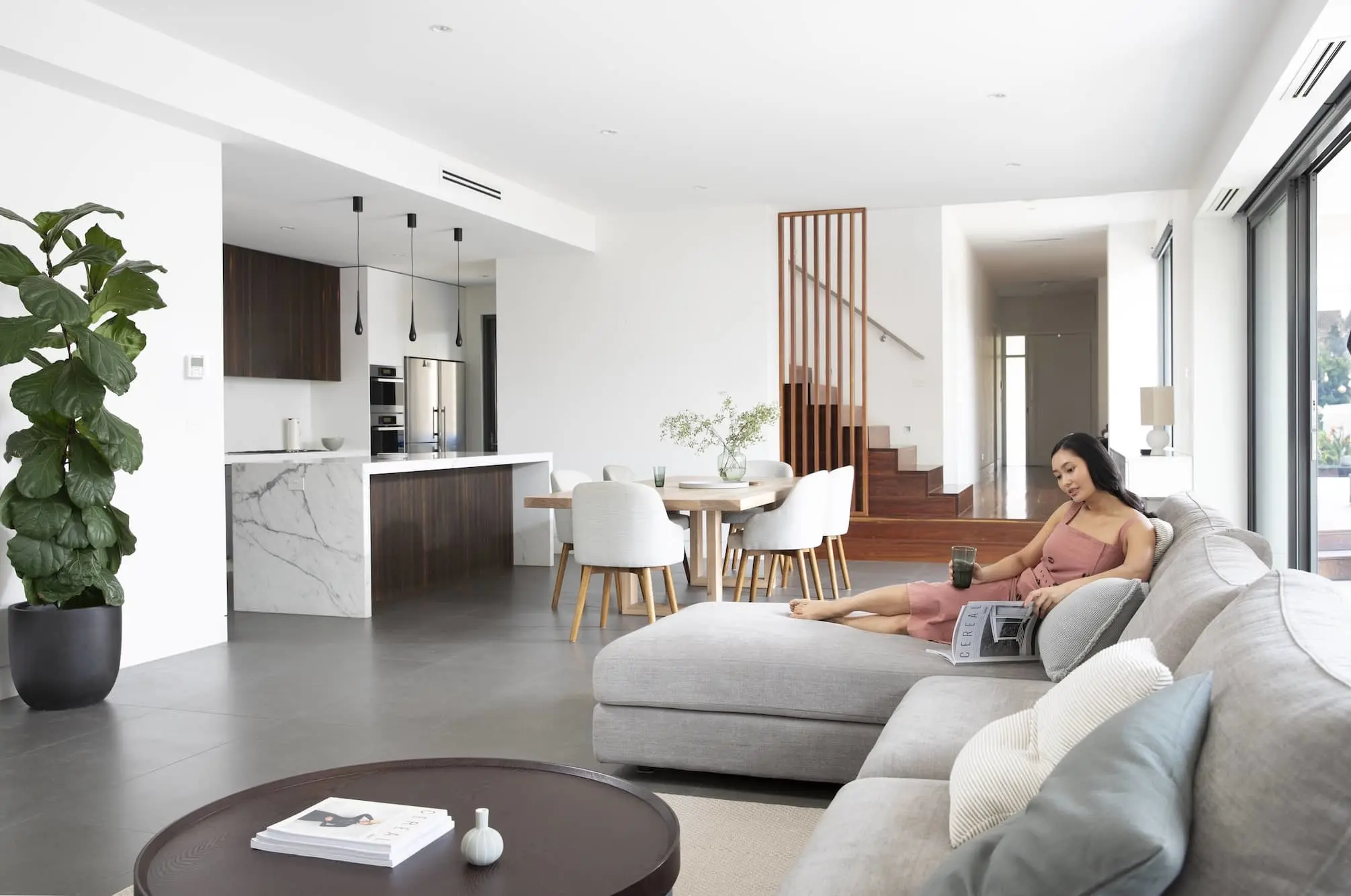 Modern open-concept living space with kitchen, dining area, and woman in pink dress relaxing on gray sectional sofa.