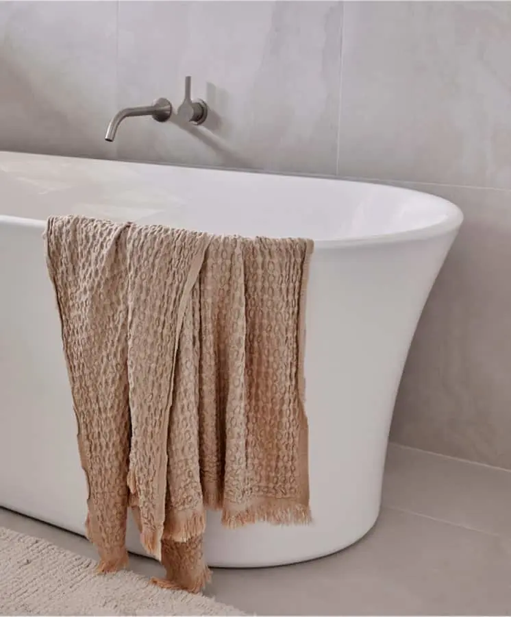 Modern white freestanding bathtub with beige textured towel draped over the edge and wall-mounted stainless steel faucet.